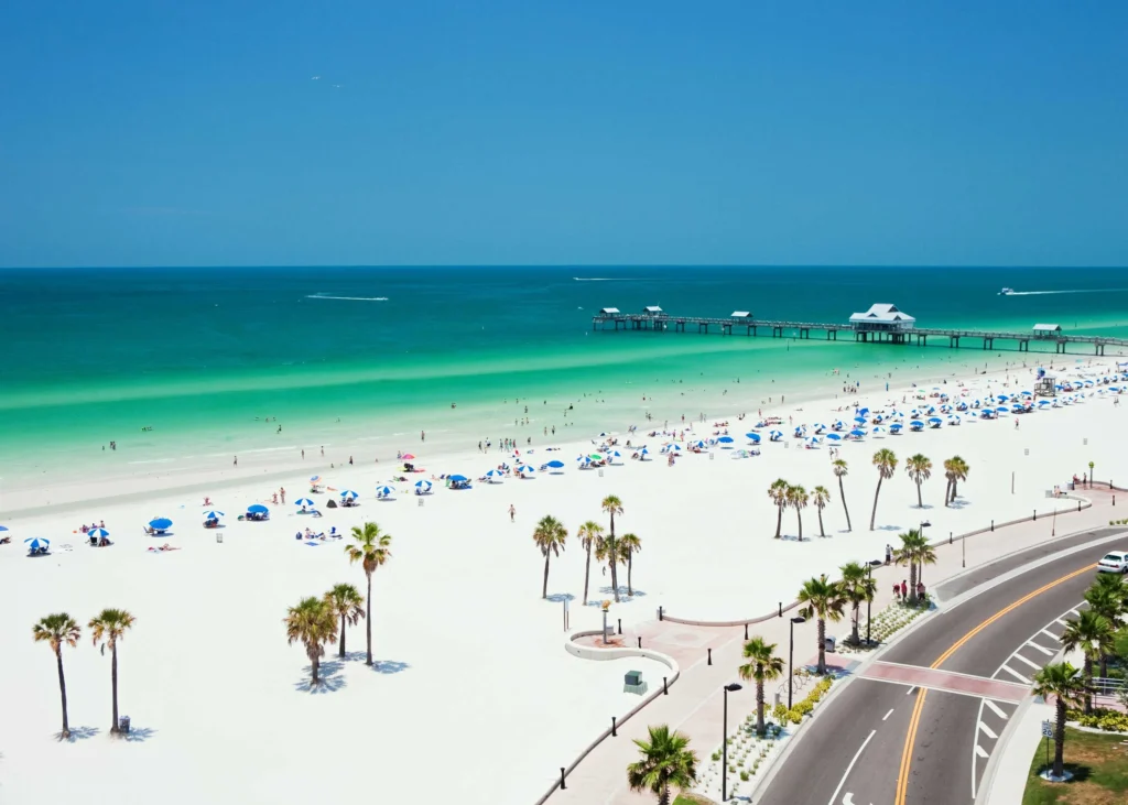 wide shot of a beach with green waters, a pier, a road, and palm trees