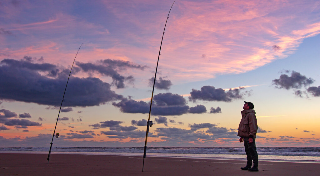 man looking at surf fishing rods on the beach at sunset