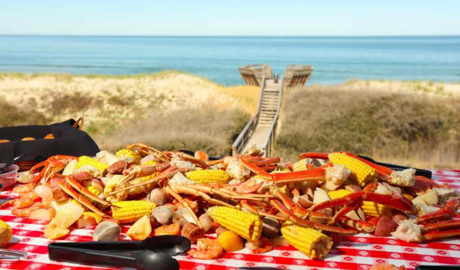 seafood boil on picnic table on the beach featuring crab legs, shrimp, corn, potatoes