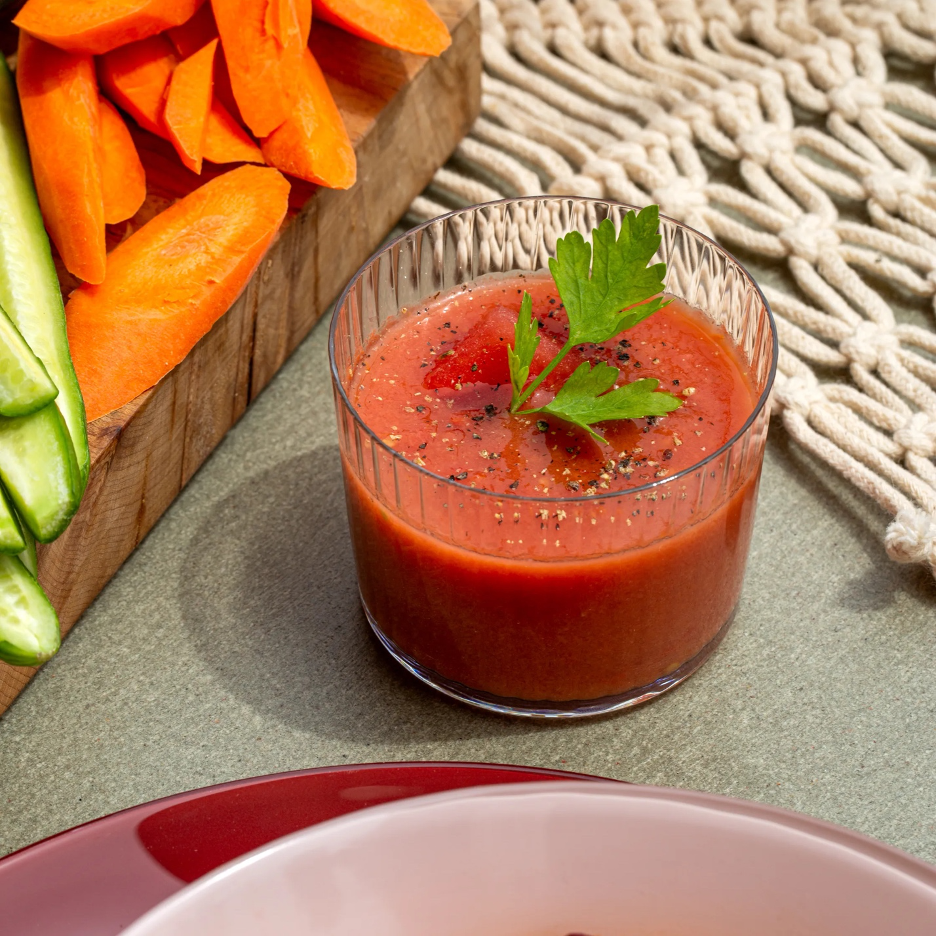 Chilled Watermelon-Tomato Gazpacho in a glass garnished with fresh herbs, and a cutting board in background with sliced carrots and jalapenos