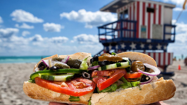 Sandwich featuring fresh tomatoes, veggies on a crusty French bread with beach and ocean background and lifeguard station on the sand