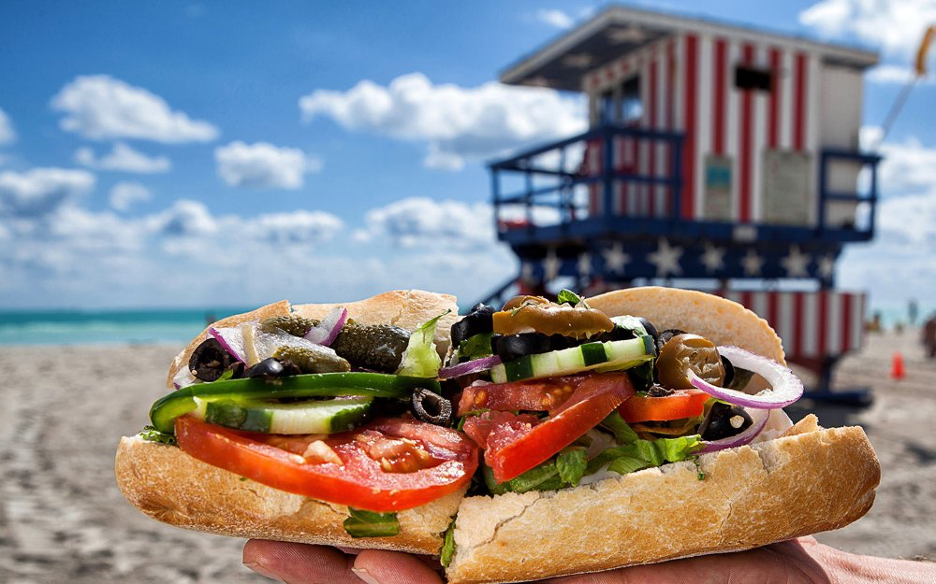 Sandwich featuring fresh tomatoes, veggies on a crusty French bread with beach and ocean background and lifeguard station on the sand