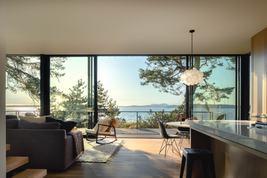 Sleek and modern living area and kitchen with outdoor deck featuring expansive coastal views, mountains, and forested landscape