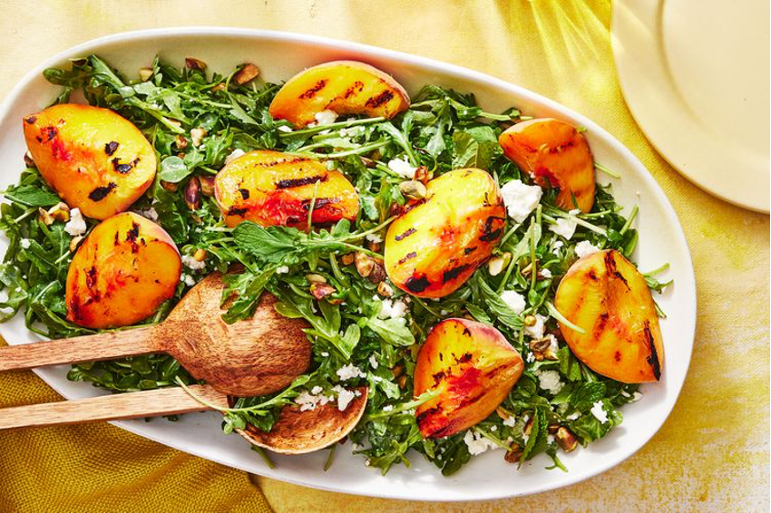 Grilled Peach & Arugula Salad with Prosciutto on a white platter with wooden serving spoons