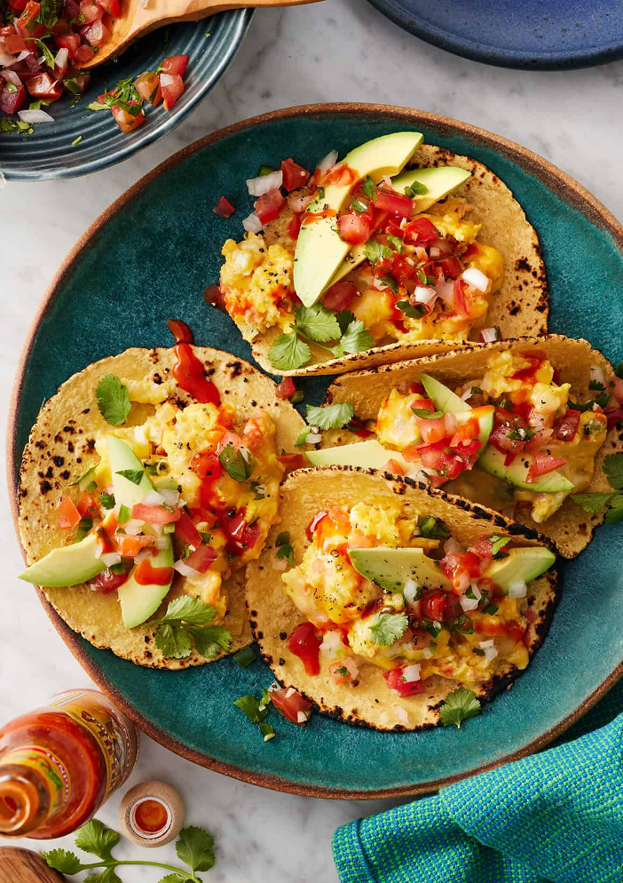 Breakfast Tacos topped with avocados, eggs, peppers, and cilantro served on a vibrant teal Mexican-style stoneware platter with hot sauce and teal napkins in foreground.