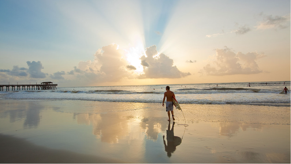 A surfer walking along the sandy shoreline of Tybee Island – Savannah, Georgia, with a fishing pier in the background