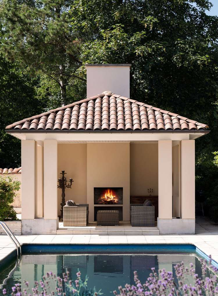 Stucco Wood-Burning Fireplace with Terra-Cotta roof Accents (SoCal) for a pool pavilion with stone pavers, outdoor seating, Spanish-style candelabras, landscaping with lavender flowering in foreground by pool