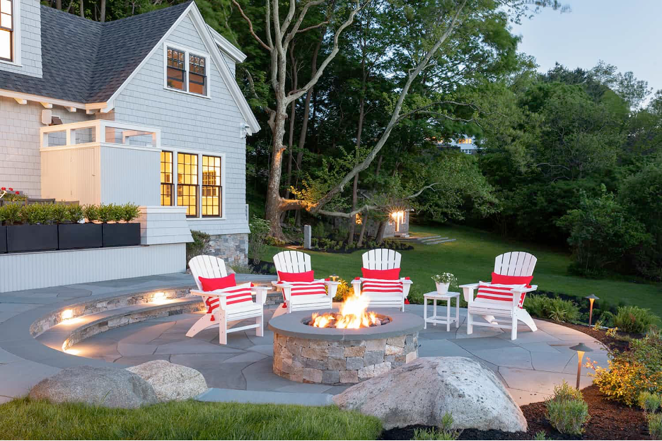 Rustic Stone Firepit with an Adirondack Vibe (Northeast Coast) for a sprawling shingled Cape Cod-style house in Massachusetts. White adirondack chairs dressed with red and white striped pillows