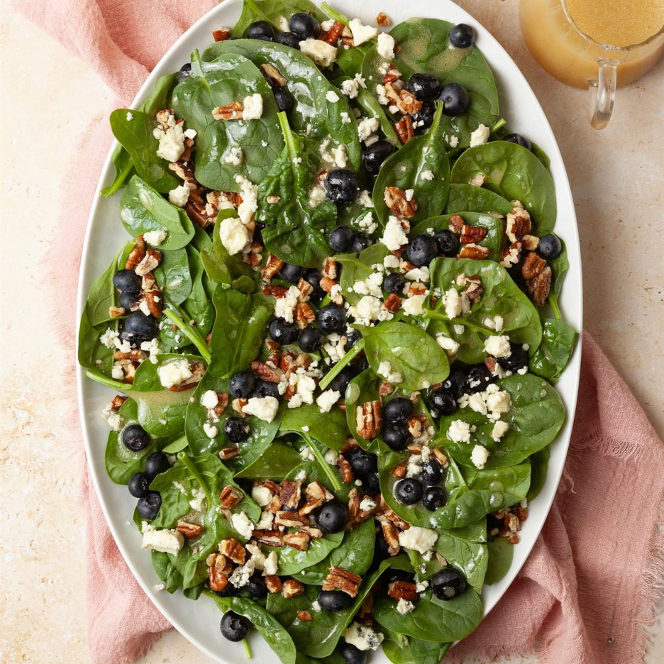 Blueberry Spinach Salad with Candied Pecans and Blue Cheese on a white oval platter with vinaigrette serving pitcher on the side