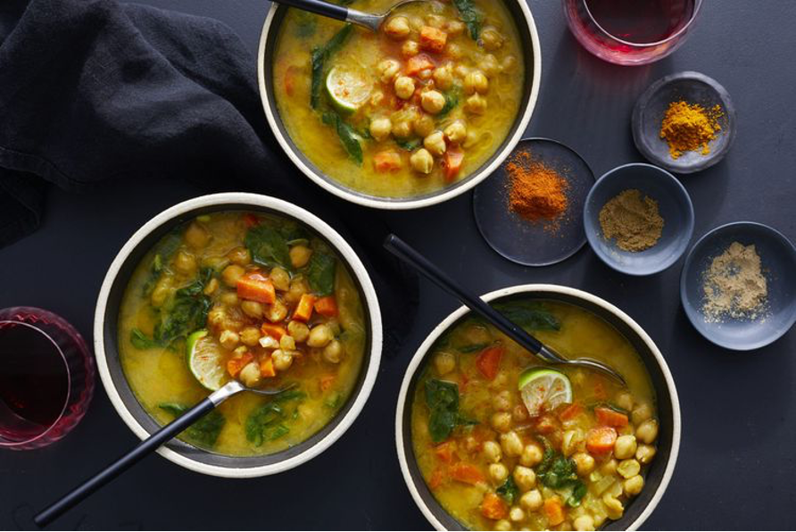 Moroccan-Style Chickpea, Carrot, and Spinach Soup in bowls with spoons on a black table with ramekins of ground spices