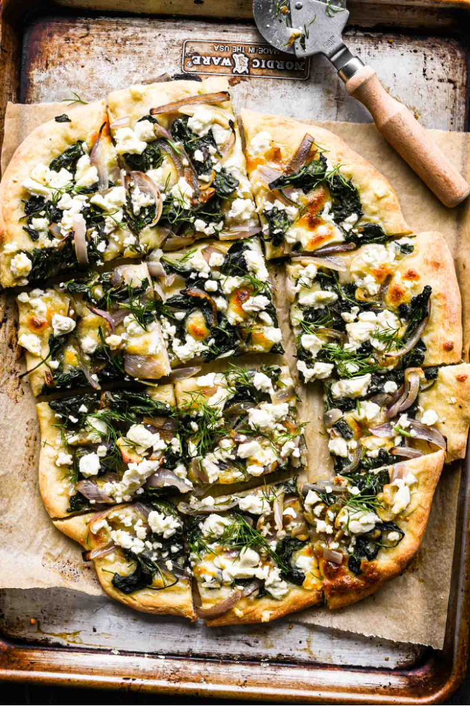 Spinach and Feta White Pizza on a baking sheet, cut into squares with a pizza cutter in background