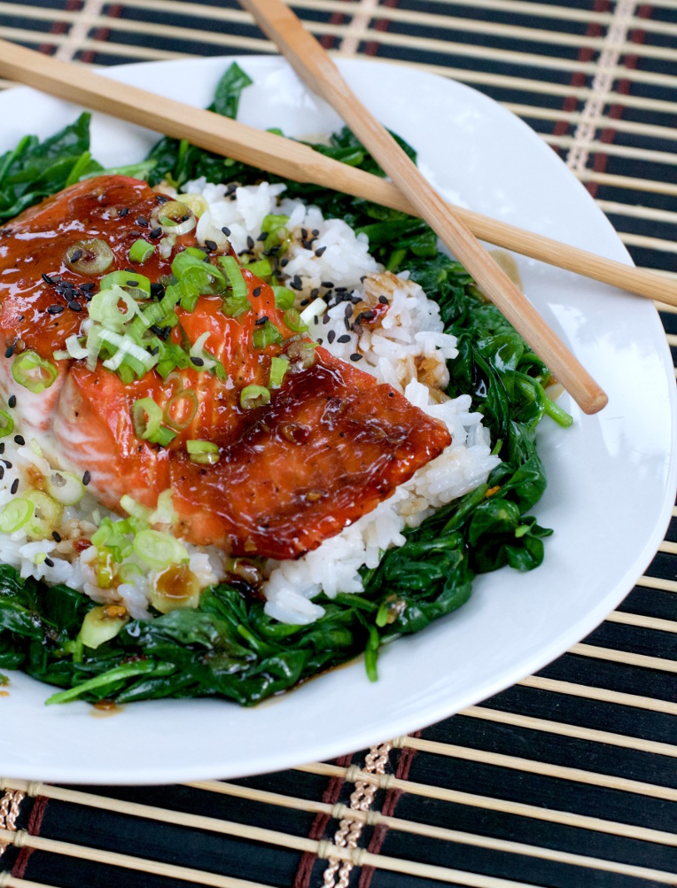 Glazed Salmon Rice Bowl Over Spinach and topped with green onions and black sesame seeds served on a white plate with chopsticks
