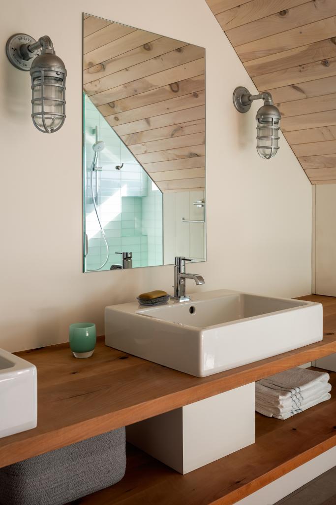 primary bath with glass-tile shower, wood-plank shelving, and countertop-mounted porcelain sinks. Light streams through a window by the shower.