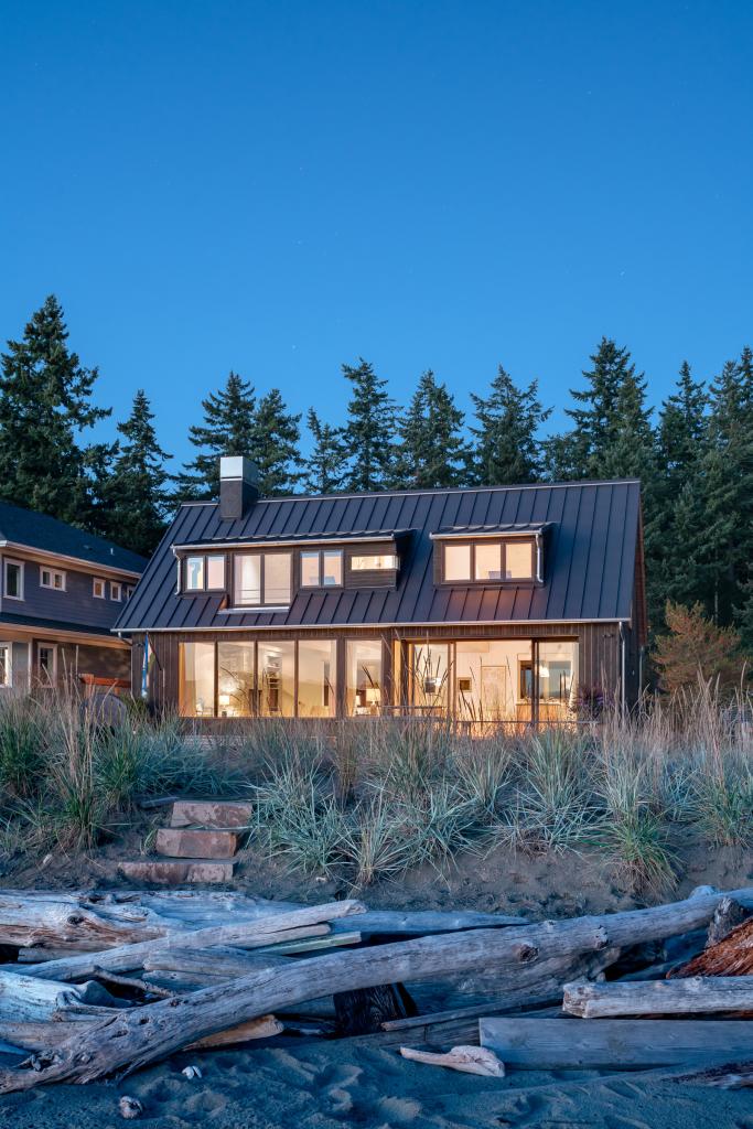 Exterior view from Mutiny Bay's sandy beach dotted with driftwood, Lit from within, the home glows and twinkles like a jewel box