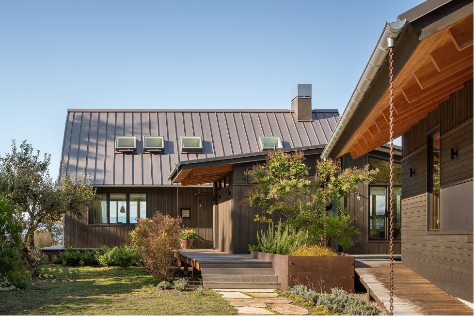 Front Exterior of Whidbey Island beach cottage in Mutiny Bay with cedar siding, a metal standing-seam roof, front gardens, a glimpse out to the beachfront and mountain views