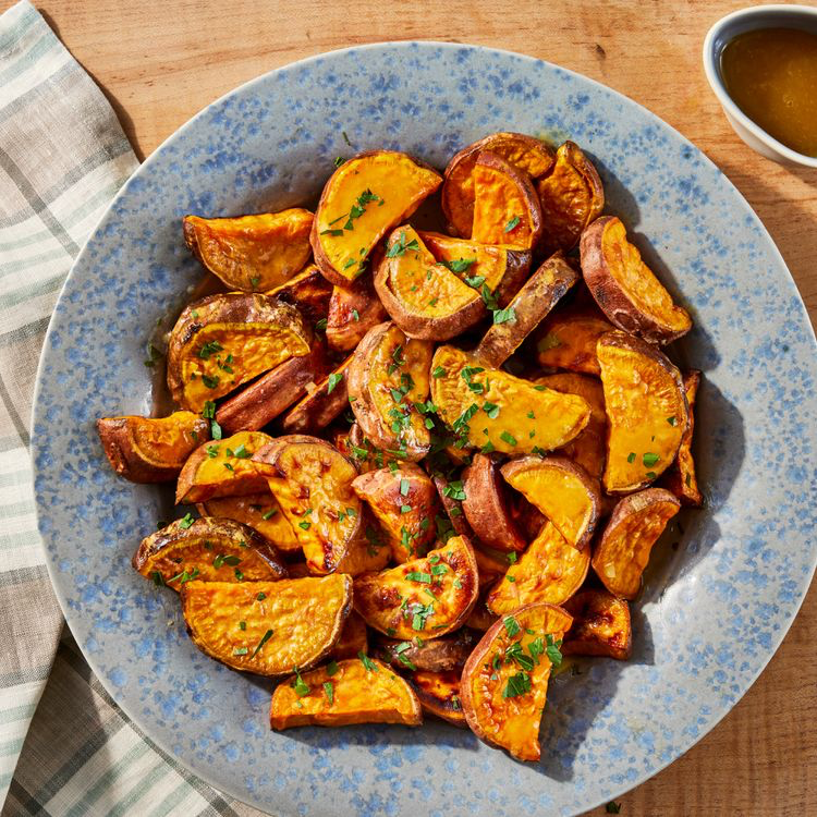 Roasted Sweet Potatoes with Citrus-Maple Glaze in a blue serving dish on wooden table with sauce and plaid linen napkin in background