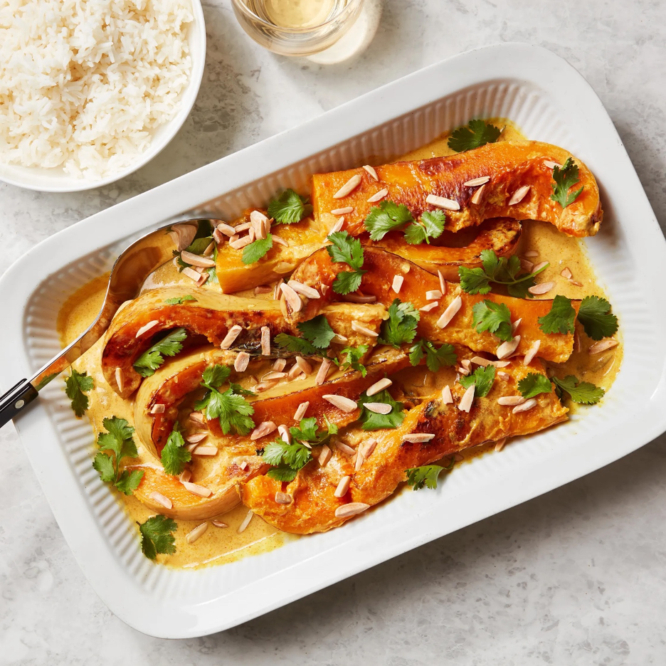 Braised Butternut Squash in Spiced Coconut Gravy in a white baking dish with serving spoon, and a bowl of rice and glass of wine in background.