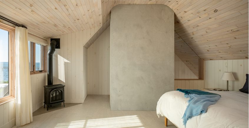 Swedish-inspired primary bedroom wrapped in bleached knotty pine with a vintage black gas stove in the corner and vast water views beyond.