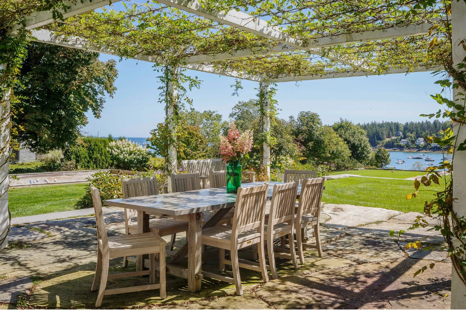coastal patio with dining area under a pergola covered with greenery, ocean background
