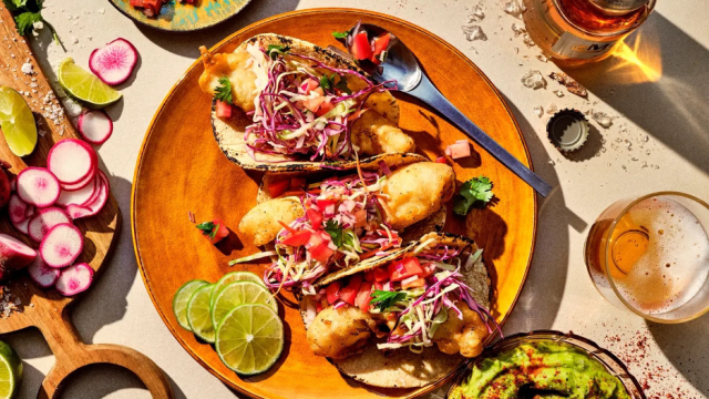 Baja fish tacos on a plate with limes, and beer and garnishes in the background