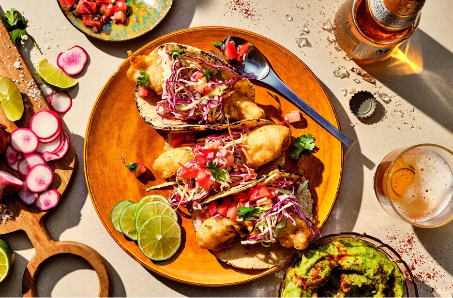 Baja fish tacos on a plate with limes, and beer and garnishes in the background