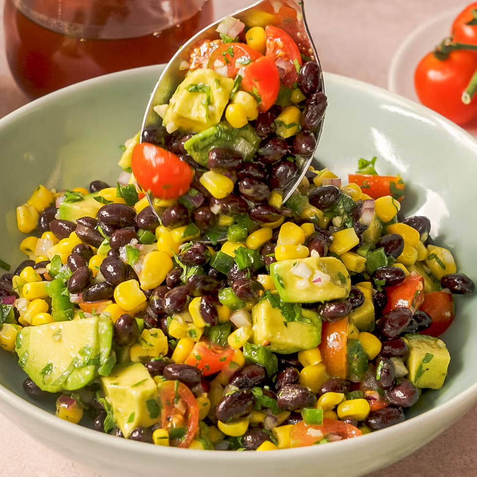 Black Bean & Corn Salad in a bowl with a serving spoon and tomatoes and honey in background